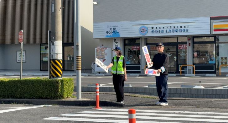 交通安全の広報活動の写真。交通安全のハンドプレートを持った人が2人横断歩道付近に立ってドライバーに呼びかけをしている様子。