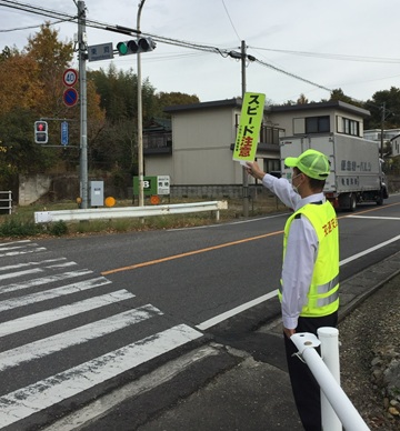 交通安全の広報活動の写真。片側1車線の道路の交差点で、黄色のベストと帽子を身につけた男性が横断歩道付近に立っている。右手には「スピード注意」のハンドプレートを持ち、ドライバーに向けて呼びかけている。