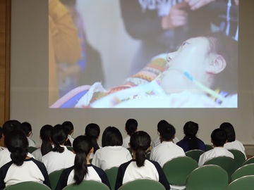 椅子に座りプロジェクターに映し出された映像を見ている生徒たちを後ろから撮影した写真。