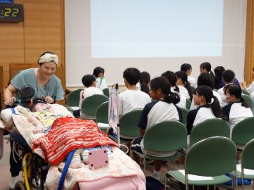 移動ベッドに横になっている歌子さんが母親の靖子さんにベッドを押してもらい会場内をぐるっと一周回っている様子の写真。生徒たちは椅子に腰かけ移動している歌子さんを見ている。