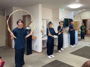 施設で「南京玉すだれ」を披露している様子の写真。利用者の方々の前で、4名の男女が円の形にした南京玉すだれを両手に持っている。