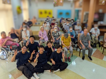 和太鼓鼓珀のメンバーが施設の方々と一緒に撮った集合写真。約30名の高齢者の方々が椅子に腰かけ、手前に鼓珀のメンバーが床に座りピースサインをしている。