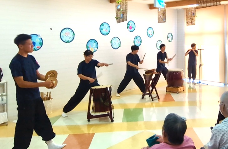 施設の方々に向けて、5人のメンバーが和太鼓、篠笛、チャッパ(両手で持って打ち合わせる小型の真鍮製シンバル)の演奏を披露している様子の写真。