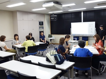 フォローアップ講座の様子の写真。養成講座で使用した部屋と同じ視聴覚室で、受講生が2グループに分かれ、テーブルをはさんで4~5人で輪になり、手話の練習をしている。