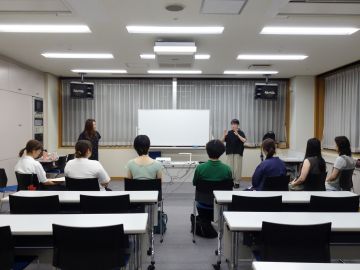 養成講座夜の部の様子の写真。手前には椅子に座った8人程の受講者が並び(後ろ姿)、奥には講師2人がホワイトボードの両側に立ち、受講者に向けて講義をしている。