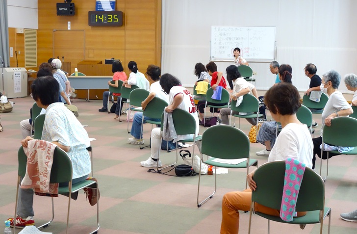 ふれあいセンターの大会議室で、椅子が2ｍ弱位の間隔でたてよこに並んでいる。参加者が椅子に横向きに座り、右足を曲げてストレッチをしている。奥の壇上には講師が同じポーズでみなさんに話しかけている。