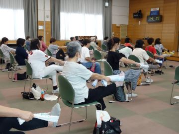 椅子に座った参加者が右足をまっすぐ体の前に上げタオルで足を引っ張っているときの写真。