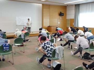椅子に座った参加者が左足の膝を伸ばし、手を腰に置いて体を前に倒している様子の写真。