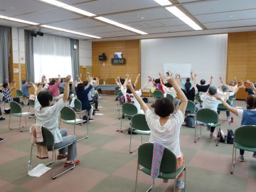 椅子に座った参加者がレクリエーションの踊りで腕を振っているときの様子の写真。