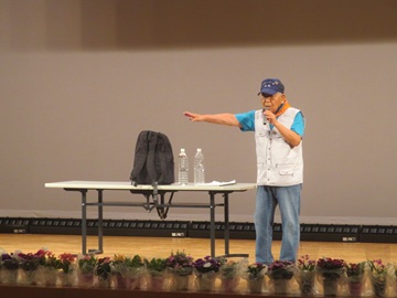 小牧防災リーダー会の写真。壇上にはキャップをかぶった70~80代の男性が一人いる。写真左側には長机があり、その上に2リットルペットボトル2本と黒のリュックサックが置いてある。左手にマイクを持ち、右手を会場の皆さんに向けている。