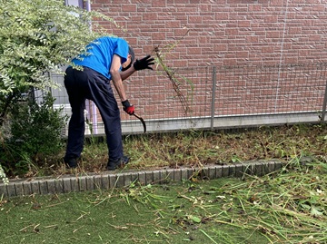 花壇の草刈りをしている様子の写真。鎌を持った男性が中腰で作業をしている。
