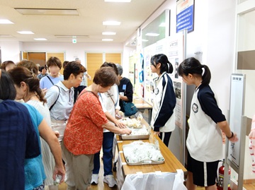 防災食の試食用のおにぎりを配っている様子の写真。長机にトレイにのったおにぎりがいくつも置いてある。机を挟んで右側にはジュニア奉仕団の生徒たち2人がいて、左側にはおにぎりをもらいに来た人や参加者が大勢いる。