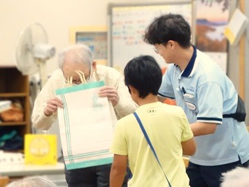 高齢者デイサービスでのプレゼント交換の様子。子どもたちからのプレゼントが入った紙袋を受け取る高齢者男性と、手前には学年代表の男の子、男性の横にはデイの職員がいる。