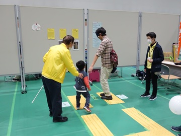 昨年の様子の写真。「ガイドヘルプ友の会」のブースでは、点字ブロックのセットが床に貼られ、来場した家族の父親がアイマスクをつけ体験している。