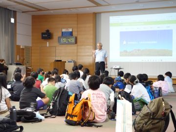 大会議室での全体説明の様子の写真。床に座っている子どもたちがおり、画像奥には児童たちに話をしているボランティアセンター所長が立っている。