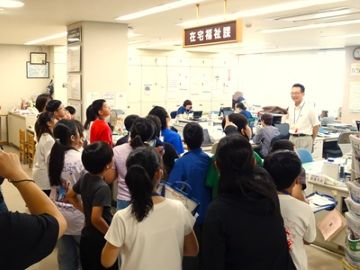 施設内見学の様子の写真。子どもたちがカウンター越しの左手に20人程立っていて、右奥には小牧市社協の次長が子どもたちに向かって笑顔で説明している。