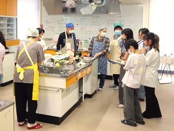 料理実習室で活動を行っていた小牧市食生活改善協議会の方たちが料理をしており、右側にいる4人の子どもたちがボランティアさんのお話を聞いている様子。
